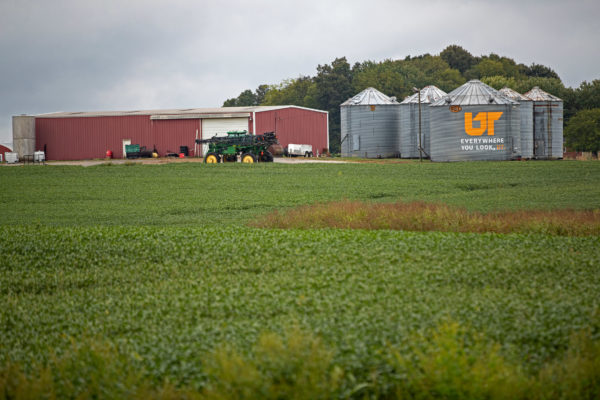 A red barn and a group of grain silos in a green agricultural field. One one of the silos has a large UT icon painted on it