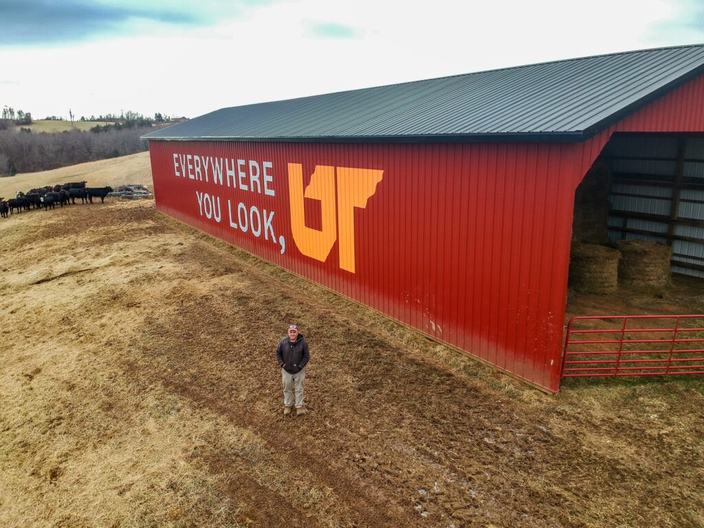 Aerial photo of red barn in Tazwell, Tennessee, showcasing Everywhere You Look, UT mural with owner/operator Whitt Shuford standing in front