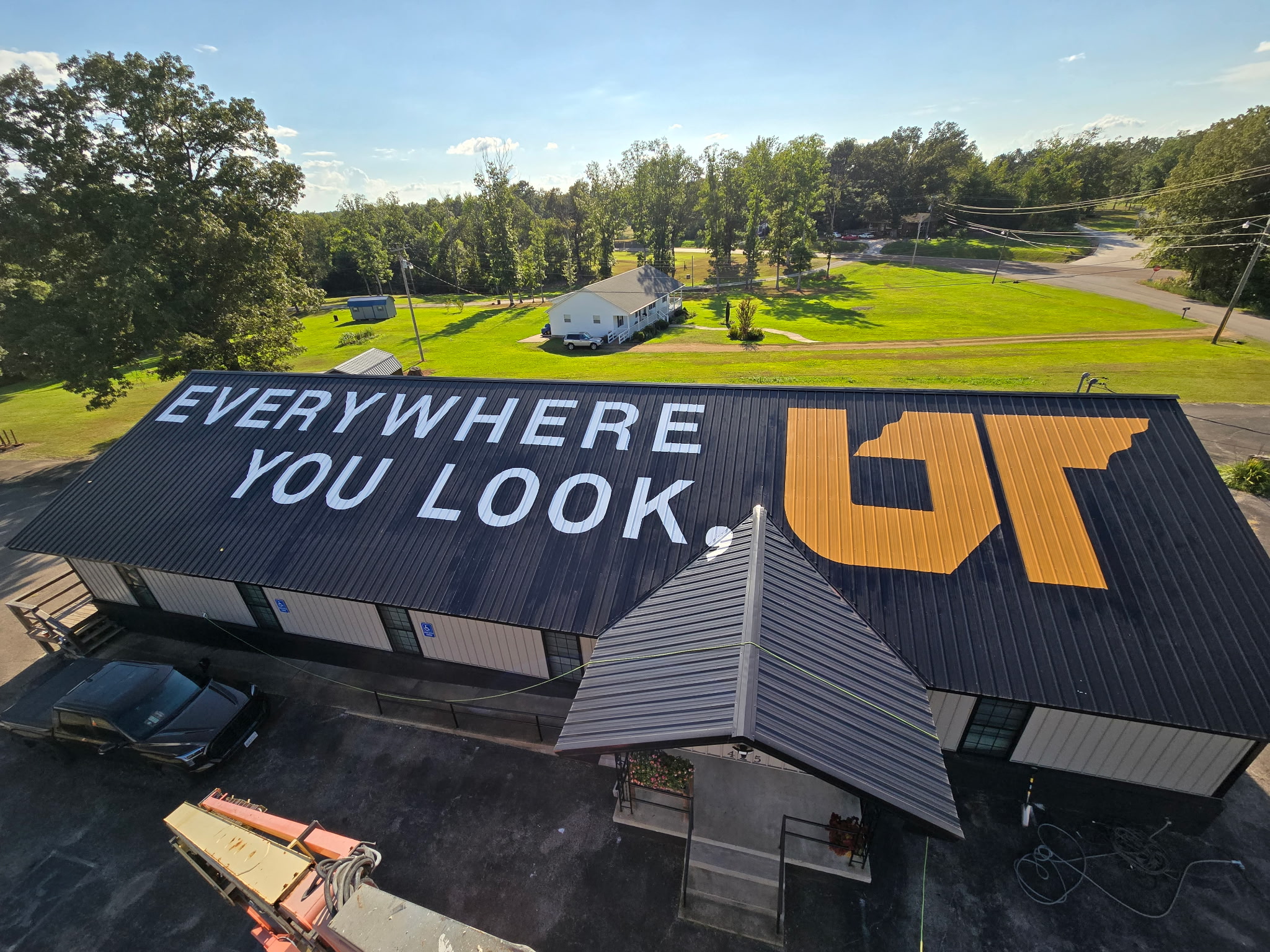 A large black metal roof featuring the words “Everywhere You Look, UT” in white and orange lettering. The mural spans the entire roof of a one-story building surrounded by trees, grass and a rural road in Decatur County, Tennessee.