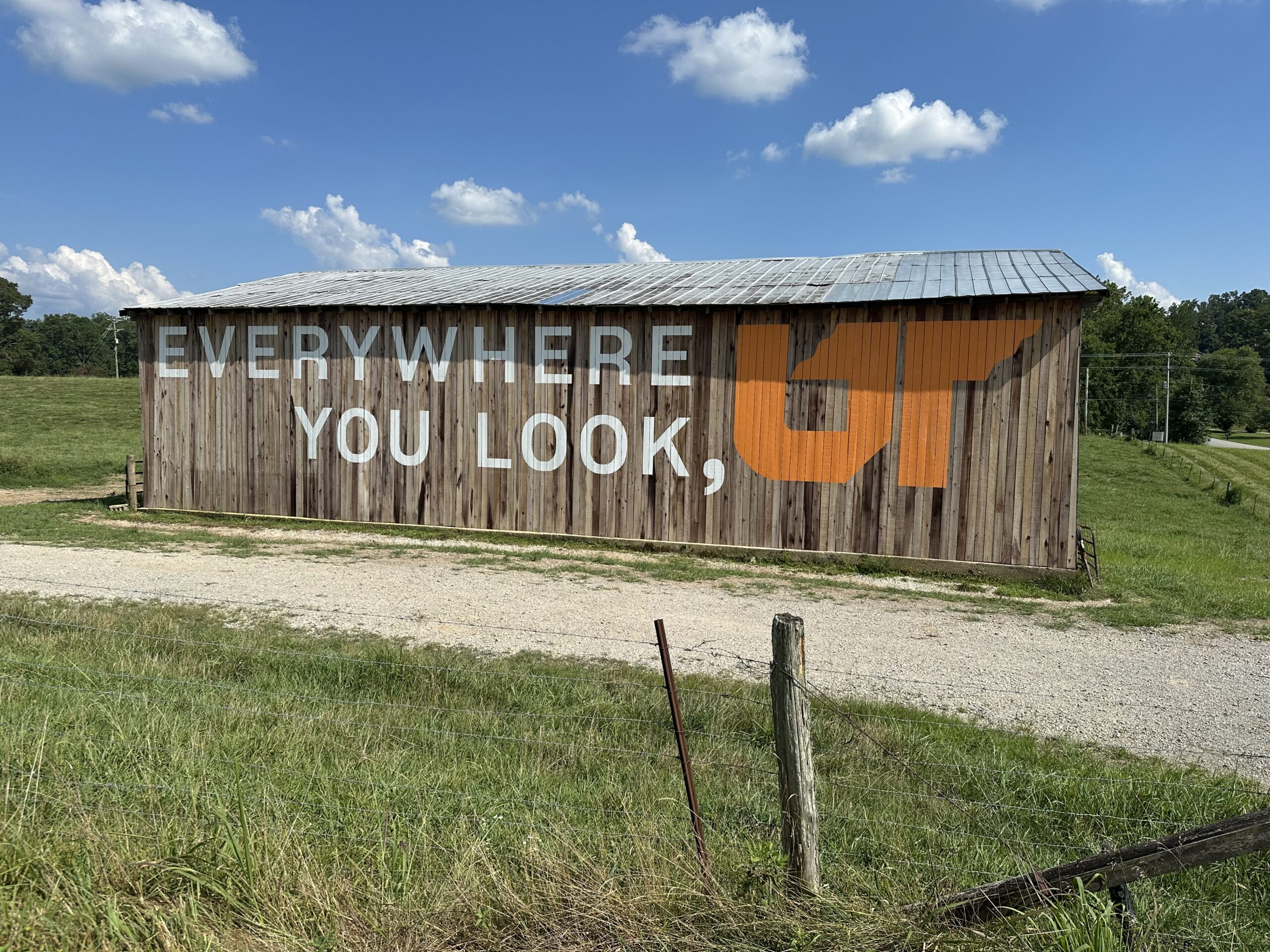 A large wooden barn in a grassy rural field painted with the words “Everywhere You Look, UT” in white and bright orange letters, part of the University of Tennessee’s mural campaign.
