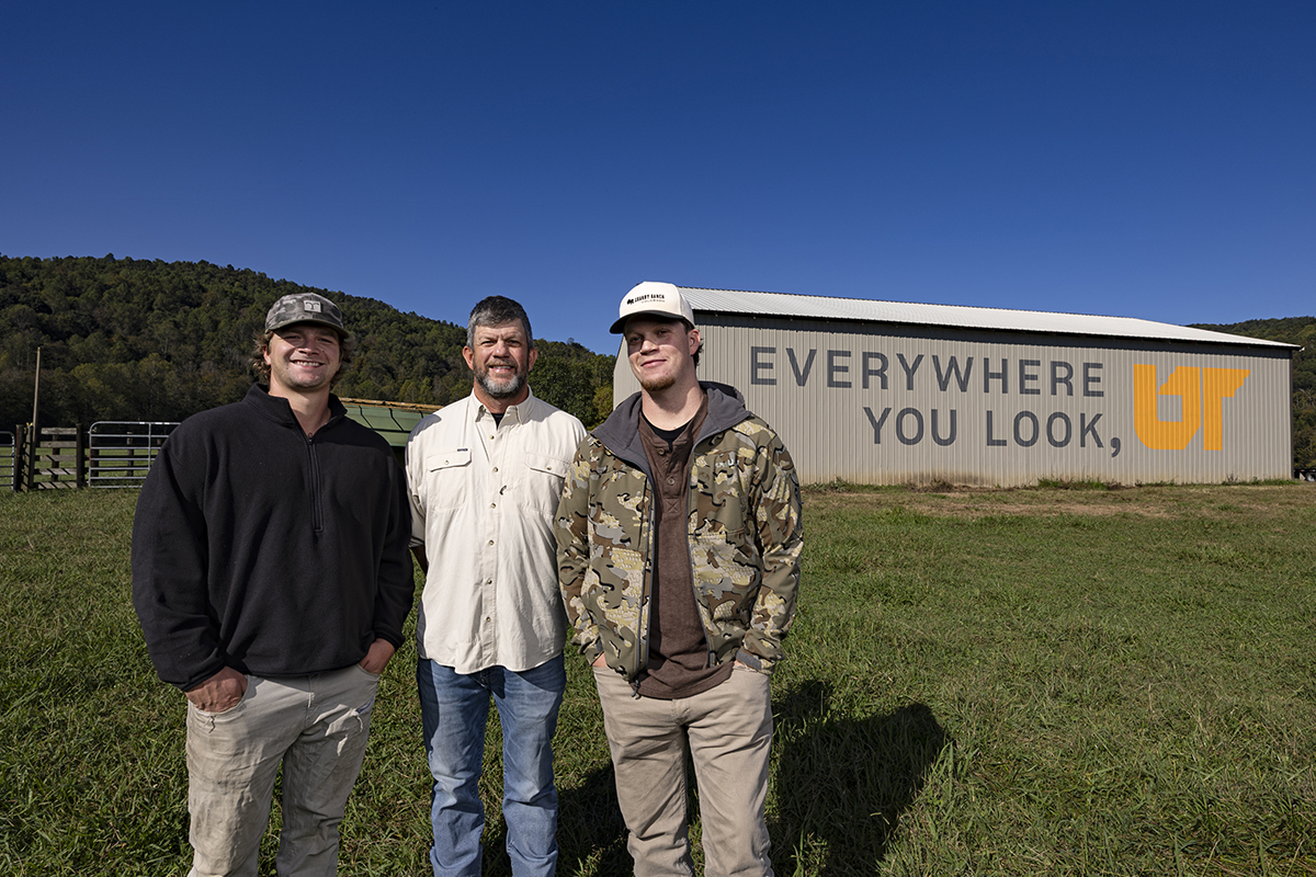 Three men standing in front of a brown barn with a painted mural.