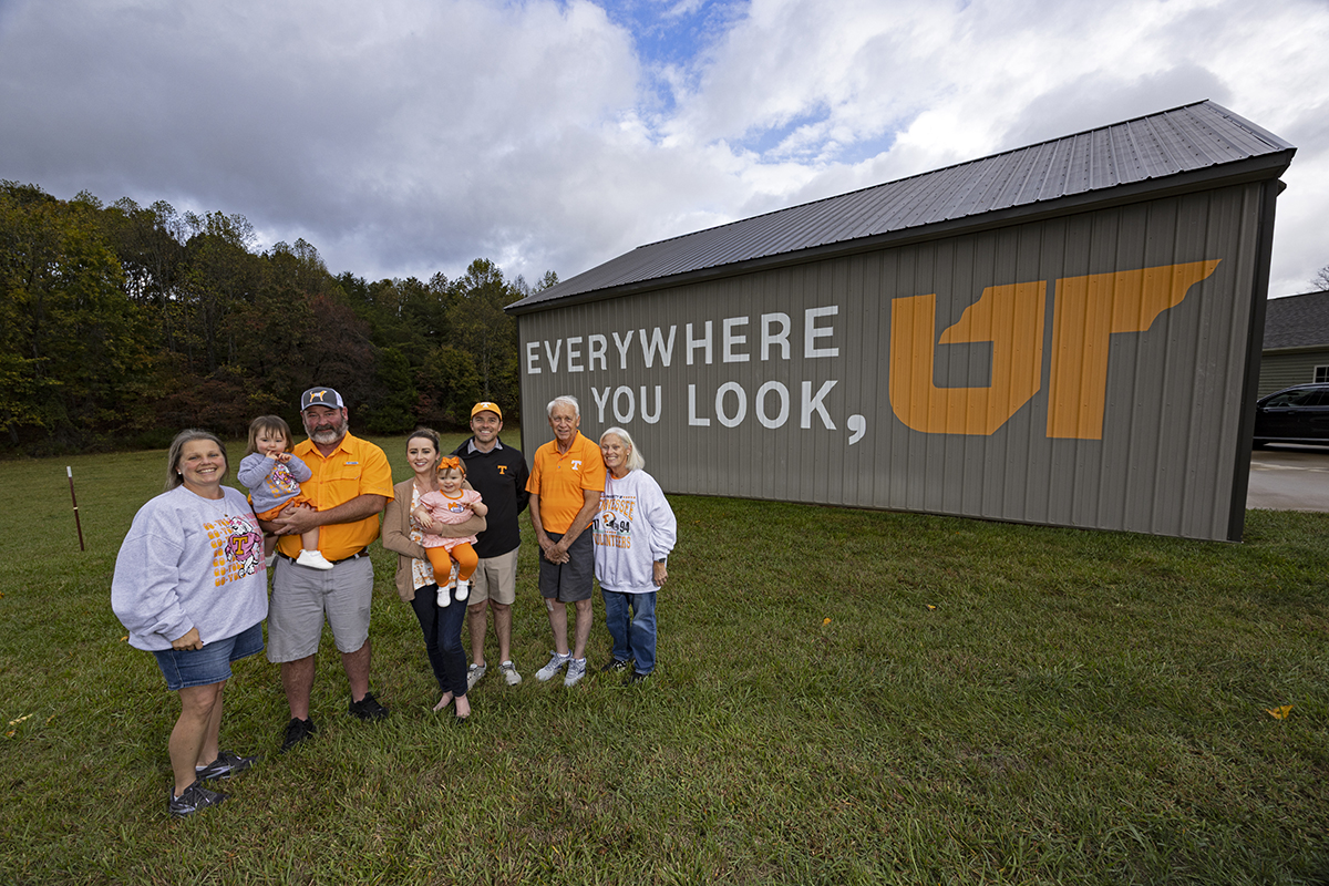 Family of eight standing in front of a brown barn with a painted mural on the side.